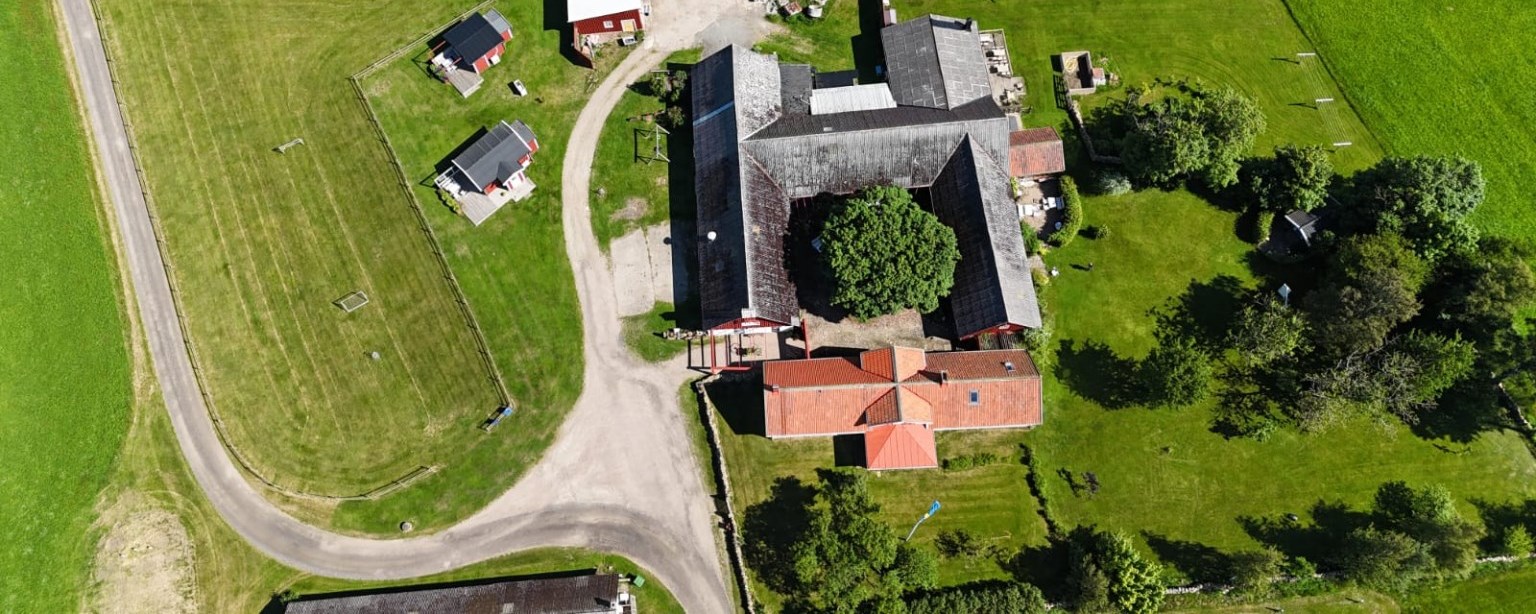 Flygbild över Rönnås Lada i Tvååker, mellan Varberg och Falkenberg, med gårdens fyrlängade byggnader, det gamla vårdträdet på innergården och det omgivande jordbrukslandskapet.