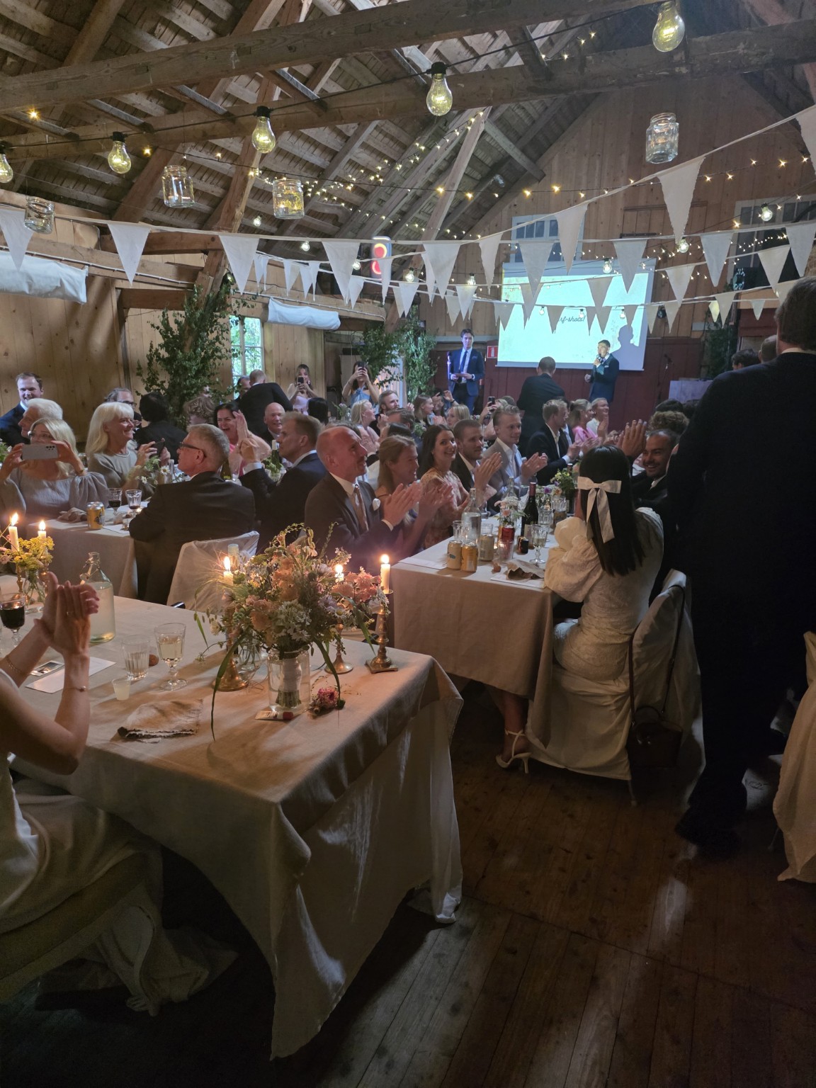 Bröllopsfest i ladan i Rönnås Lada, full av applåderande gäster med ljusslingor och vimplar i taket.
