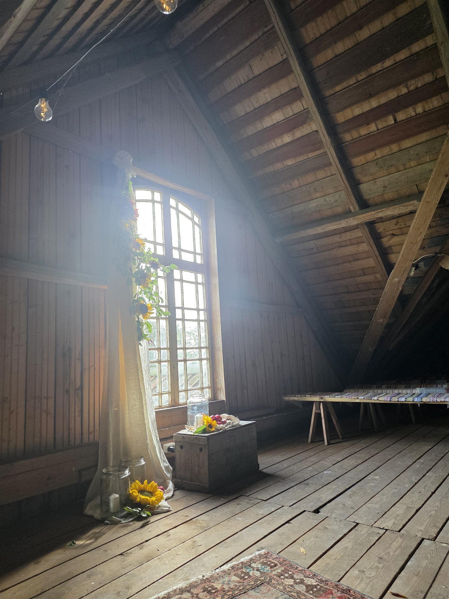 Loftet i ladan i Rönnås Lada, där solljuset strömmar in genom det stora fönstret och vigselplatsen är smyckad med solrosor och tyg.