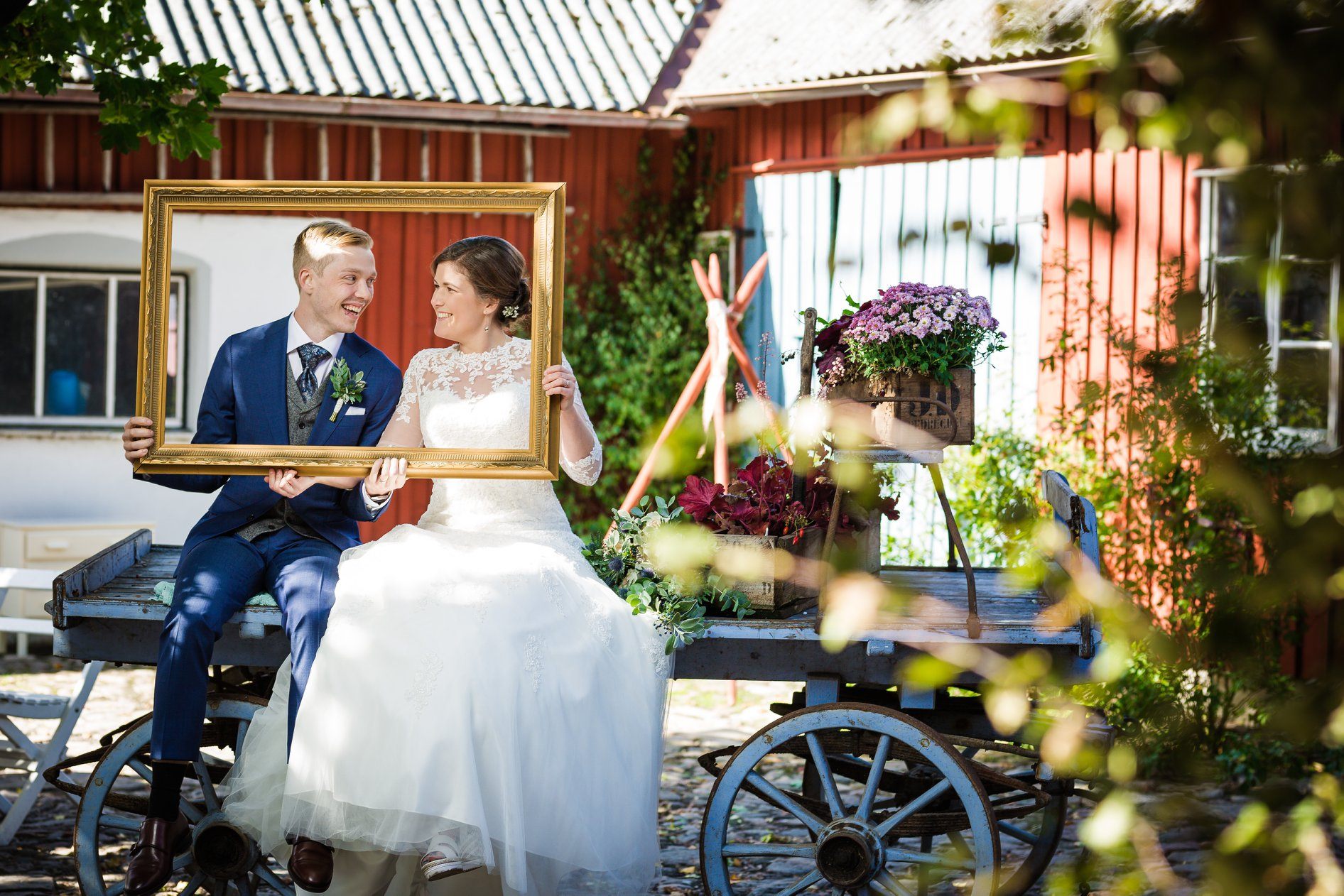 Brudpar sitter på en gammal vagn framför de faluröda husen och håller upp en guldram.