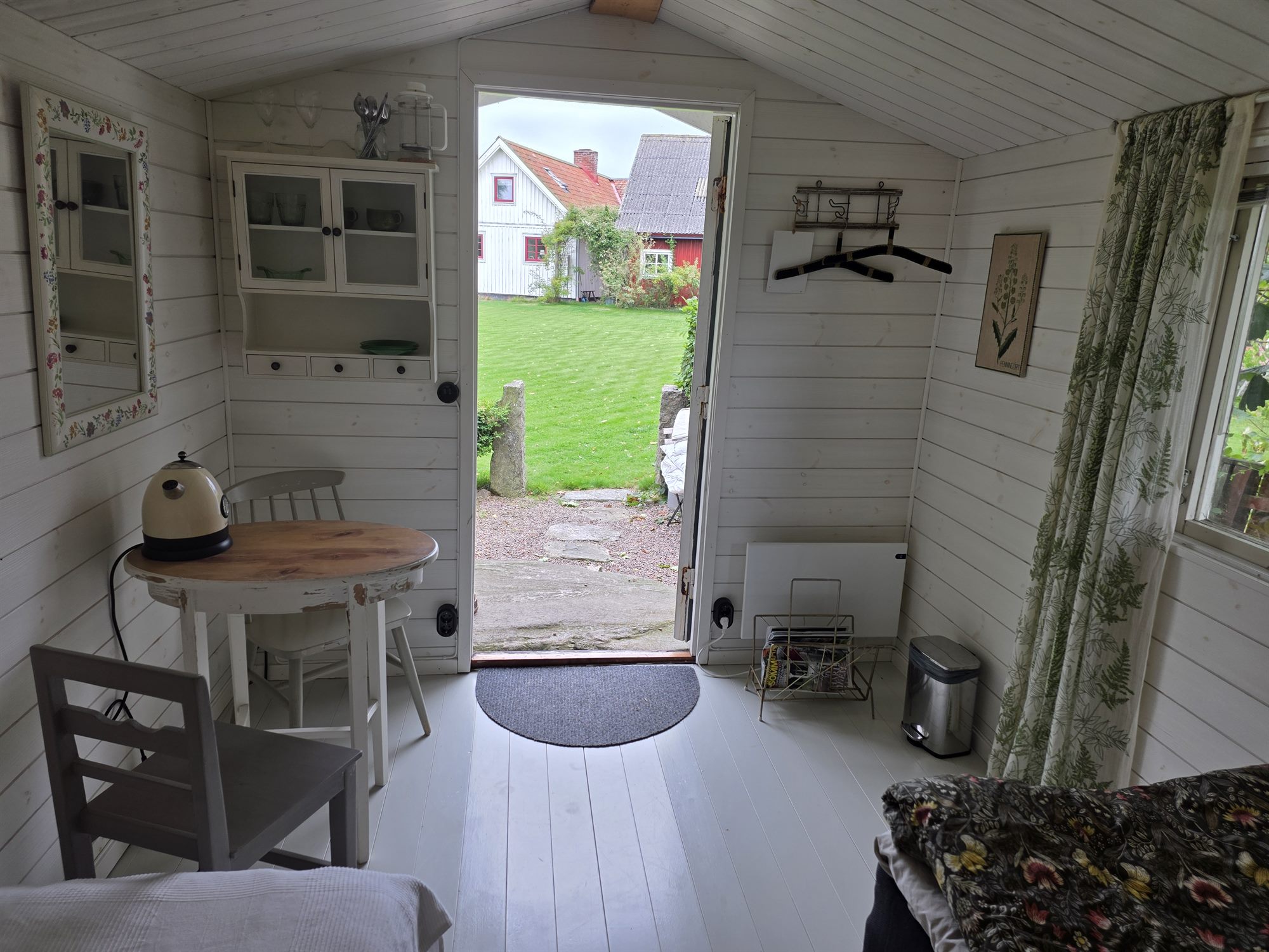 Interiör i Trädgårdsstugan med öppen dörr ut mot gräsmattan och stenläggningen.