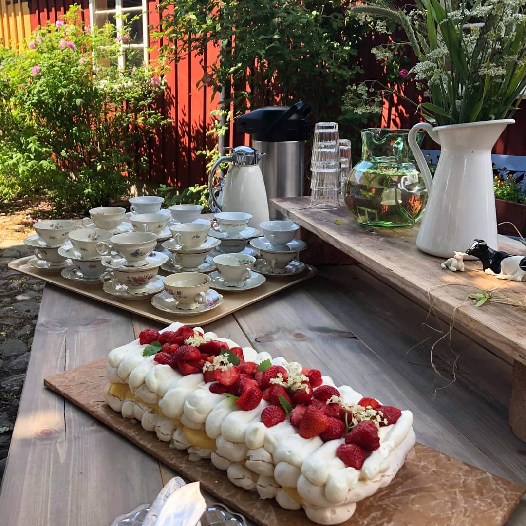 Jordgubbstårta med maränger dukad utomhus, med uppradade kaffekoppar och en termoskanna intill.